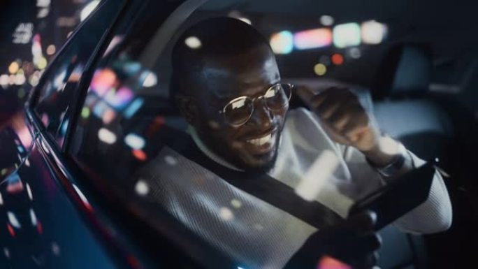 时髦的戴眼镜的黑人在晚上坐出租车的后座上下班。英俊的男性在智能手机上玩电子游戏，而在城市街道上的一辆