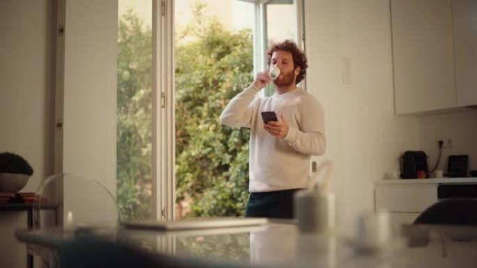 英俊的成年男子，姜黄色卷发，使用智能手机，站在家里的客厅。男性喜欢喝一杯浓缩咖啡，查看社交媒体，在线