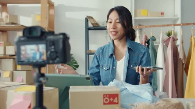 年轻的亚洲女士时装设计师使用手机接收订单，并在商店在线展示服装录制视频直播。