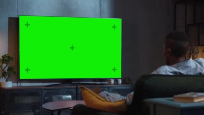 黑人坐在家里的沙发上看绿色模拟屏幕电视。男流晚间电影。肩膀上的景色。娱乐概念