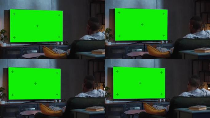 黑人坐在家里的沙发上看绿色模拟屏幕电视。男流晚间电影。肩膀上的景色。娱乐概念