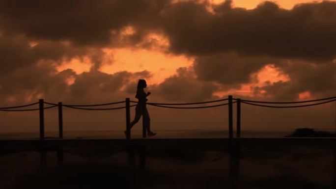 在深橙色的日落中，无法辨认的女性在海滩上的木板路上奔跑的照片
