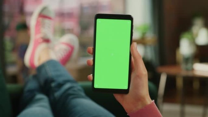 女性手握着带有绿屏模拟显示的智能手机。女性正在家里的沙发上放松，在移动设备上观看视频和阅读社交媒体帖