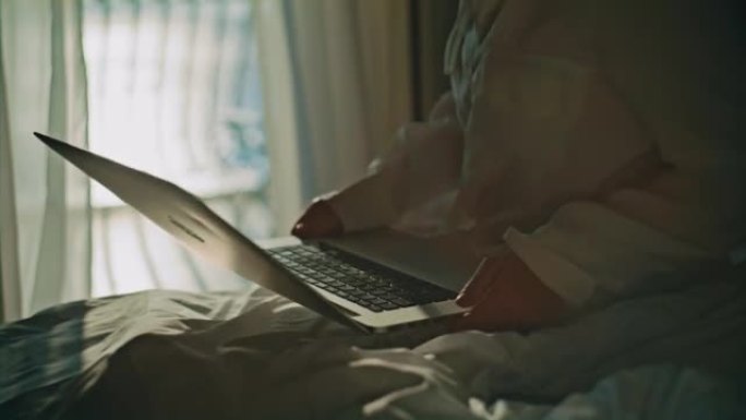 金发女人从她房间的酒店阳台上走，坐在床上，开始在笔记本电脑上工作