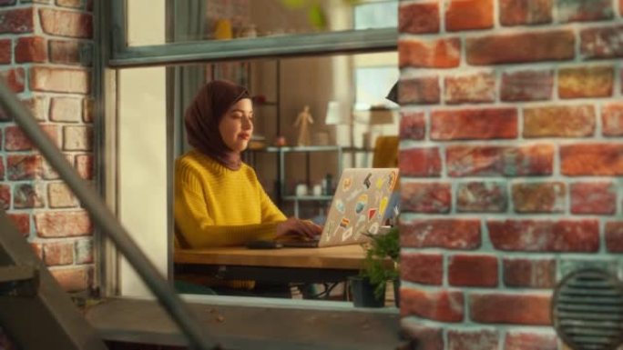年轻女性戴着头巾，使用笔记本电脑，坐在她公寓的客厅。穆斯林女商人检查社交媒体，微笑，在家回复电子邮件