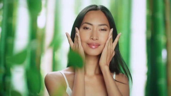 美丽的亚洲女人轻轻涂抹面霜的肖像。年轻的成年女性使用科学先进的天然化妆品护肤品使皮肤柔软光滑。绿色森