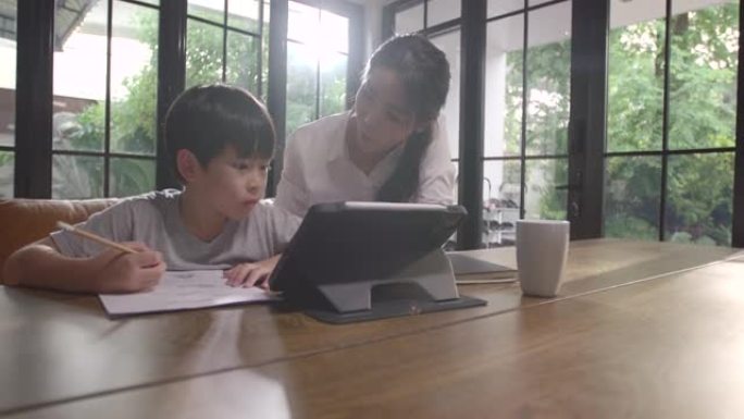 一个亚洲男孩正坐在家里专心地在线学习，记下他通过平板电脑学到的知识，母亲在他身边教书。由于Covid