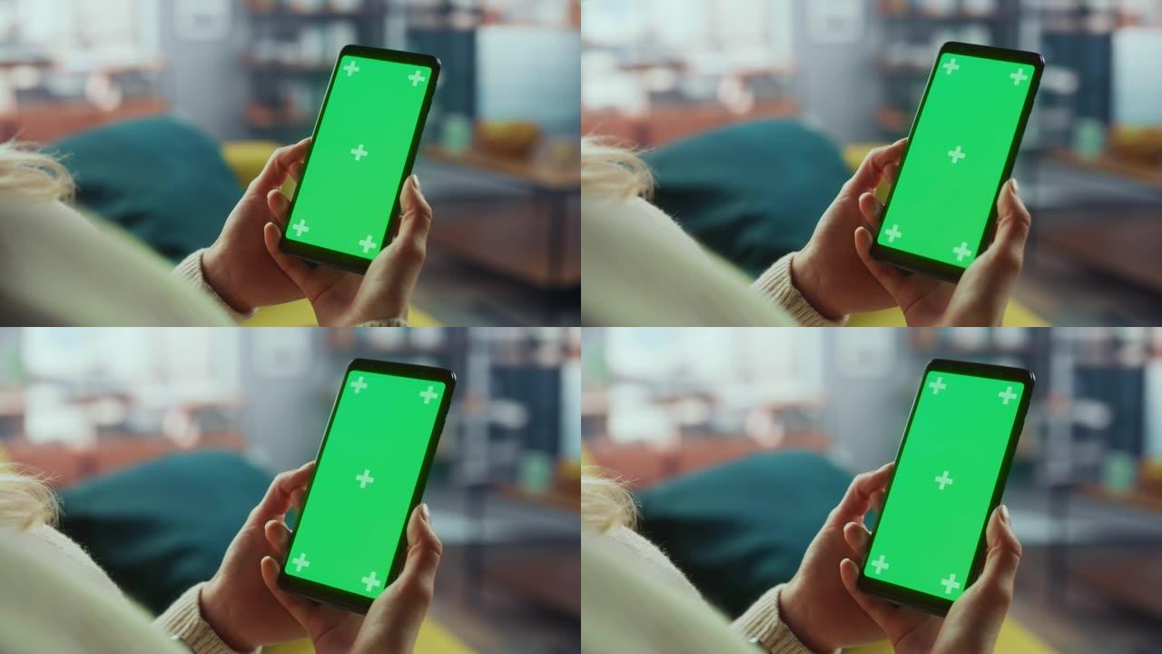 躺在沙发上时，在家里的客厅使用带有绿屏模拟显示的智能手机的真实女性的特写镜头。她正在浏览互联网并在社