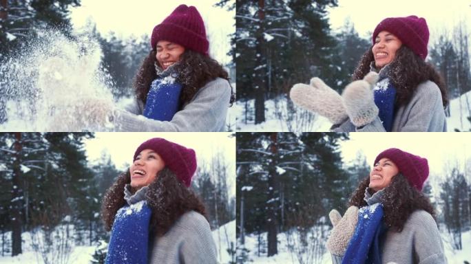 冬季乐趣。美丽的年轻快乐的混血女人穿着温暖的手套在雪地上吹着雪，微笑着在白雪皑皑的森林慢动作。