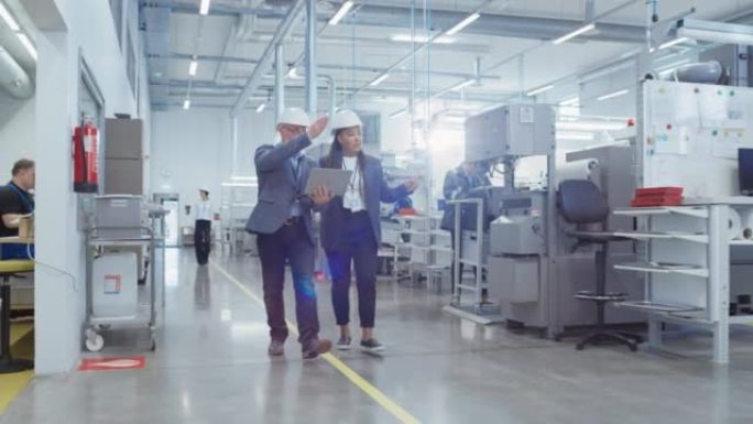 两名专业重工业工程师在工厂戴着安全帽。步行和讨论工业机器设施，在笔记本电脑上工作。非裔美国人经理和技