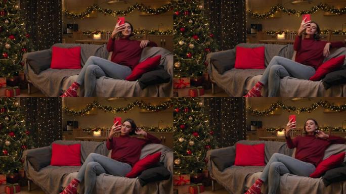 一个年轻漂亮的女人正在她的红色智能手机上自拍，坐在一个房间里的沙发上，房间里有圣诞灯和一棵装饰好的圣