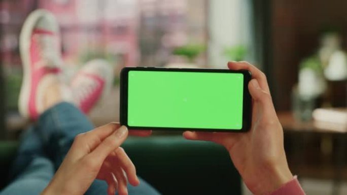 女人水平拿着智能手机，绿屏模拟显示。女性正在家里的沙发上放松，在移动设备上观看视频和阅读社交媒体帖子