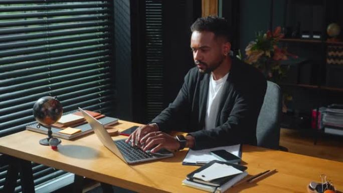 年轻的专注于严肃的非裔美国商人使用笔记本电脑在办公室的桌子上工作，拿起电话接听电话。