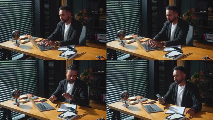 年轻的专注于严肃的非裔美国商人使用笔记本电脑在办公室的桌子上工作，拿起电话接听电话。