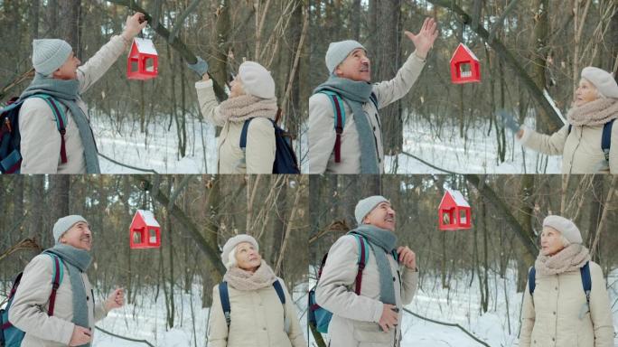 高级夫妇在树枝上悬挂鸟屋，在冬季公园散步时聊天