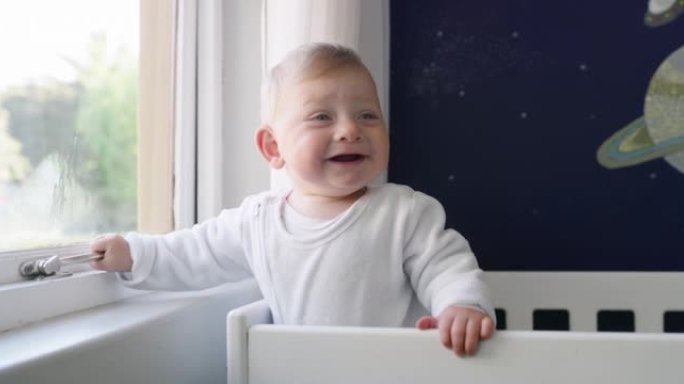 4k视频片段，一个可爱的男婴站在家里的婴儿床里在窗户上玩耍