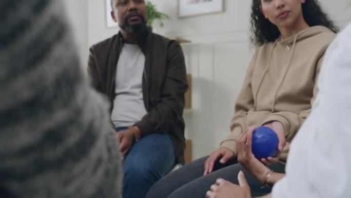 一个由不同的人组成的支持小组的特写镜头，他们坐在一个圈子里，在小组治疗期间传递一个蓝色的球并交谈。各