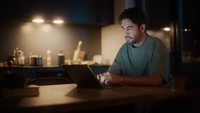 英俊的地道拉丁男人坐在舒适的厨房里的桌子旁，晚上在家使用笔记本电脑。男性成年人在浏览互联网时思考，在