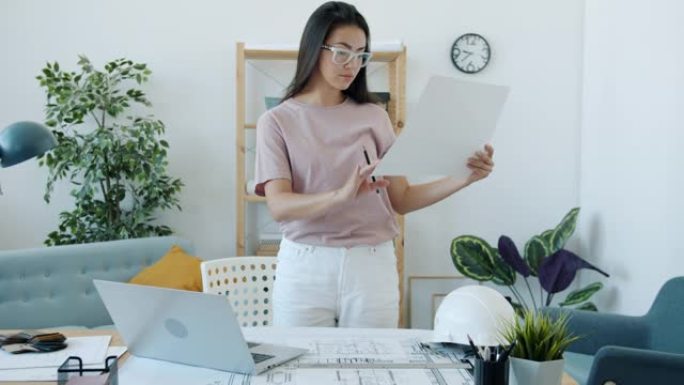 年轻的亚洲建筑师抱着蓝图思考，独自在家使用笔记本电脑