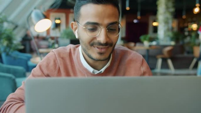 快乐的阿拉伯男人的肖像在咖啡馆里戴着空气舱和使用笔记本电脑聊天，而人们在背景中行走