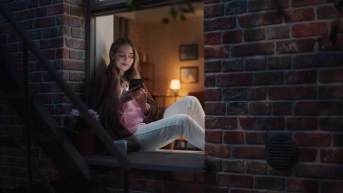 一位美丽的白人妇女晚上坐在窗台上，穿着羊毛毯子，正在使用智能手机。年轻女性在浏览在线约会应用程序中的