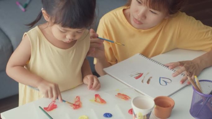 快乐快乐的亚洲家庭妈妈教蹒跚学步的女孩油漆陶瓷锅在客厅的桌子上放松。