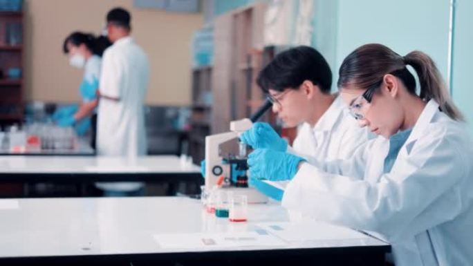 在研究实验室中，穿着白大褂的工业科学家和工程师组成的多元化团队与显微镜一起工作