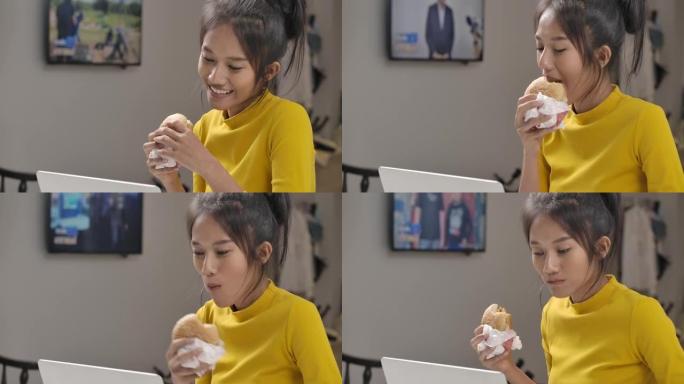 年轻美丽的亚洲女人在工作或使用笔记本电脑在线学习时吃汉堡。检疫期间，快乐的女孩穿着黄色休闲服在家里微