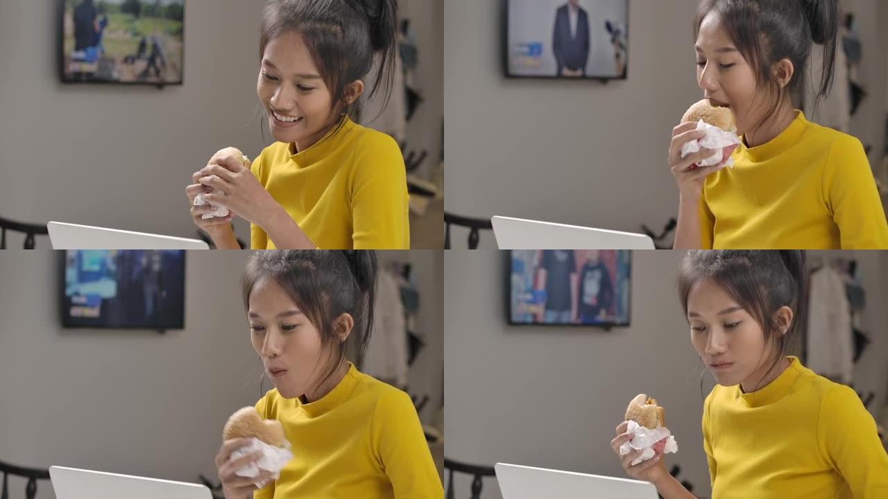 年轻美丽的亚洲女人在工作或使用笔记本电脑在线学习时吃汉堡。检疫期间，快乐的女孩穿着黄色休闲服在家里微