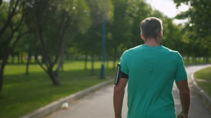 穿着绿松石t恤的跑步者的后背，手臂上有一个跑步的步伐监视器，他清晨正在公园里散步
