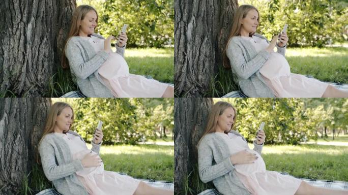 在城市公园的树下使用智能手机拍摄快乐的未来母亲的肖像