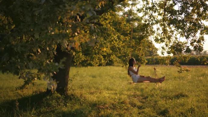 慢动作，手持一个年轻女孩摇摆的镜头，坐在一个悬挂在阳光充足的草地上的大树枝上的绳索秋千上