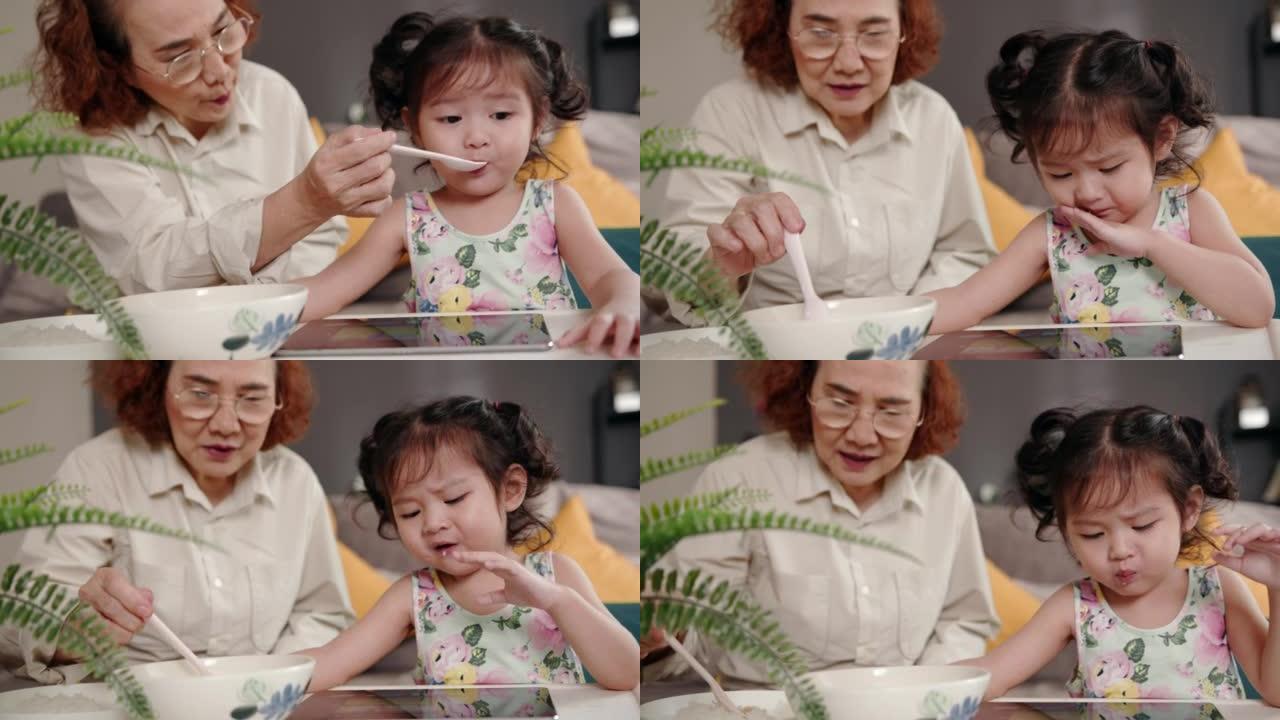 祖母一边给可爱的蹒跚学步的孙女喂饭，一边在家看数字平板电脑上的卡通。选择性饮食，对新食物的恐惧