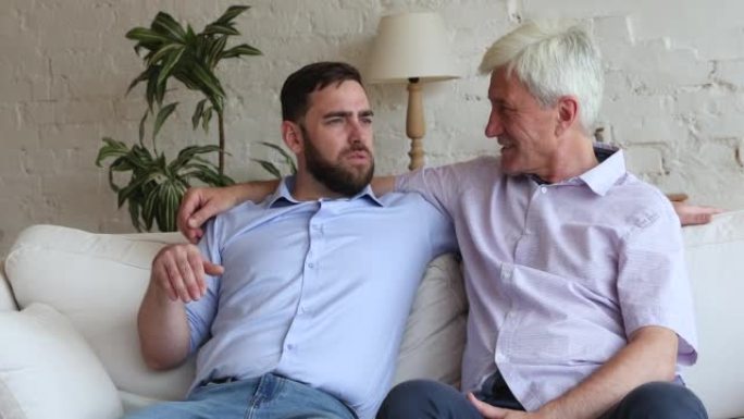 千禧一代男子拥抱他的资深父亲坐在沙发上说话