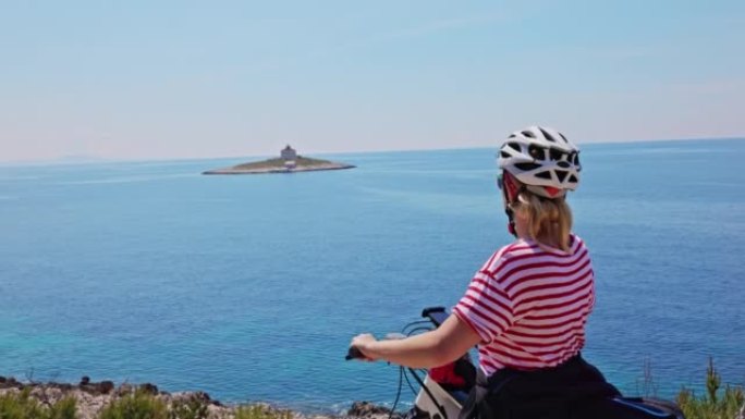 骑自行车的女人到达赫瓦尔岛的海岸，休息一下，看着一个上面有一所房子的小岛