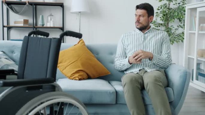 可怜的残疾人坐在沙发上叹息的慢动作肖像，轮椅坐在最前面