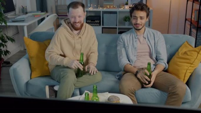 快乐的白人和中东年轻人晚上在家看电视笑着聊天喝啤酒