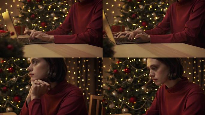 一位穿着红色毛衣的年轻美女正坐在带圣诞灯的厨房的桌子旁，在笔记本电脑上打字。