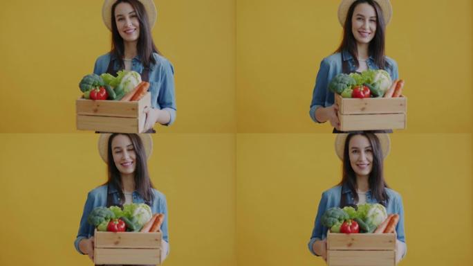 年轻女子的肖像拿着装有新鲜蔬菜的木箱，在黄色背景上微笑