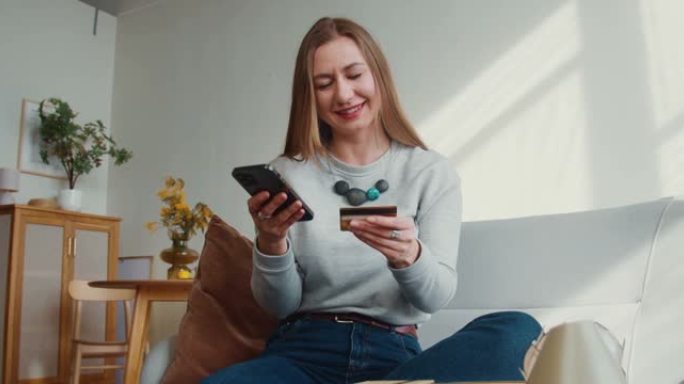 购买在线概念。美丽快乐的年轻微笑的女人在家里使用智能手机应用程序输入信用卡号码。