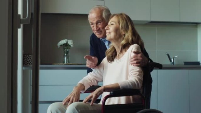 微笑的成熟丈夫拥抱轮椅上的残疾妻子的电影镜头，是在家中透过窗户看时永远永恒的爱的标志。生活观念，家庭