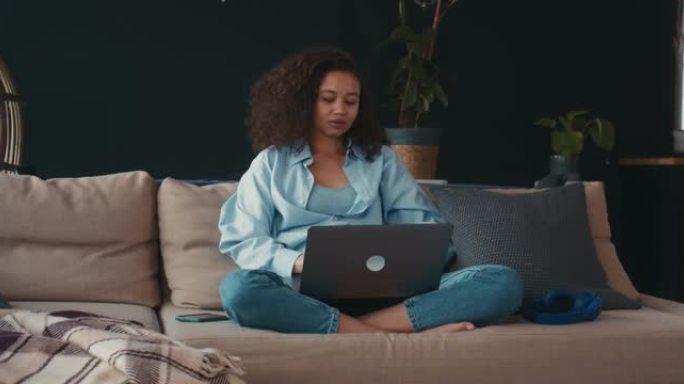家庭生活。美丽专注的年轻混血女人，卷发，在家使用笔记本电脑和智能手机工作。