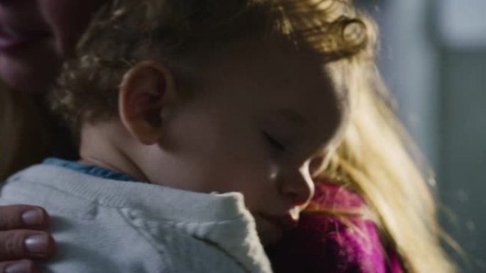 年轻的快乐和平母亲的电影镜头正在拥抱和唱歌摇篮曲她的蹒跚学步的男婴，同时睡在家里的厨房里。甜梦、童年