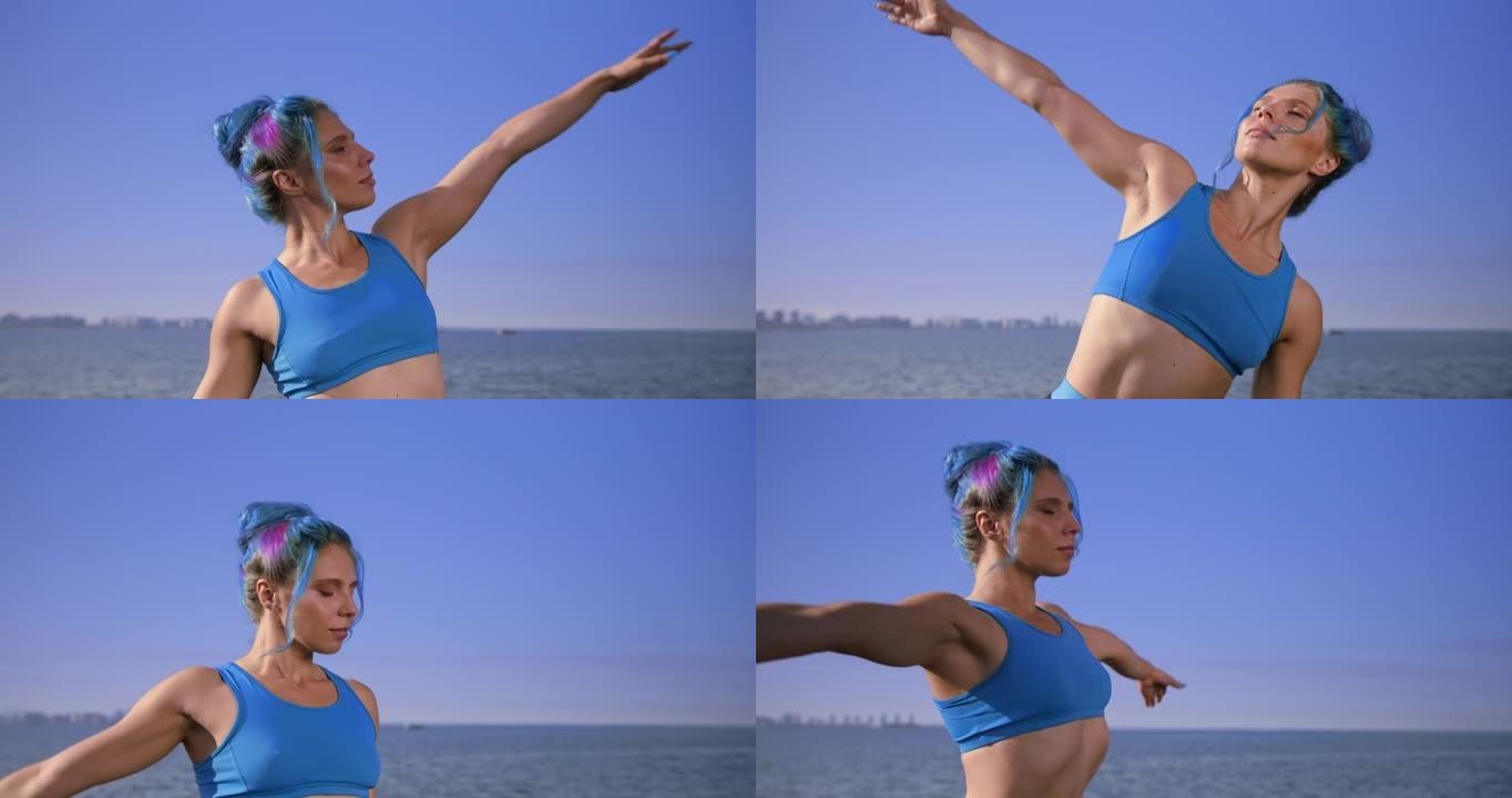 迷人的健身瑜伽老师，绿松石色的运动上衣中的绿松石色头发站在天空和大海的背景下，展示了肩膀的一些伸展动