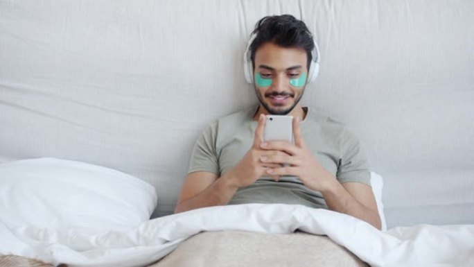阿拉伯帅哥戴着眼罩，戴着智能手机，戴着耳机在床上听音乐