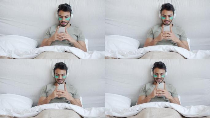 阿拉伯帅哥戴着眼罩，戴着智能手机，戴着耳机在床上听音乐