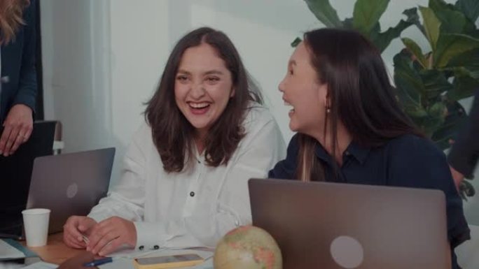 工作场所多样性两个快乐的多民族女性业务经理坐在一起在办公室的桌子上开会慢动作。