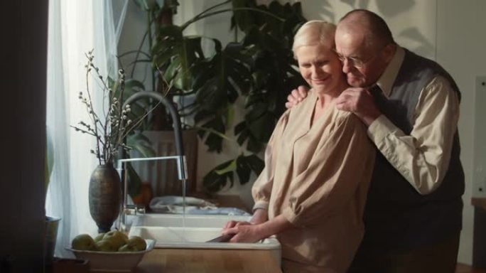 爱情和婚姻。快乐甜蜜浪漫60年代的高级丈夫拥抱美丽的妻子洗碗，在舒适的厨房微笑。