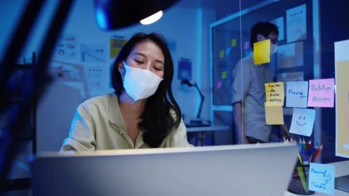 快乐亚洲女商人戴着医用口罩，在新的正常情况下保持社交距离，以预防病毒，同时在办公室晚上工作时使用笔记