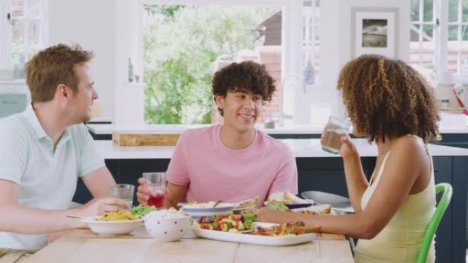 十几岁的儿子与多种族的父母坐在家里厨房的桌子旁，一起吃健康的家庭餐-慢动作拍摄